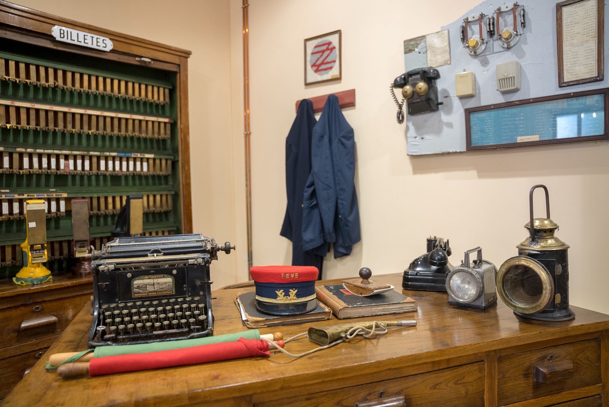 Escritorio con máquina de escribir Continental, gorra FEVE, banderines rojo y verde, instrumento de aviso, libros, secante, teléfono, linterna y farol; armario de billetes y comunicaciones detrás.