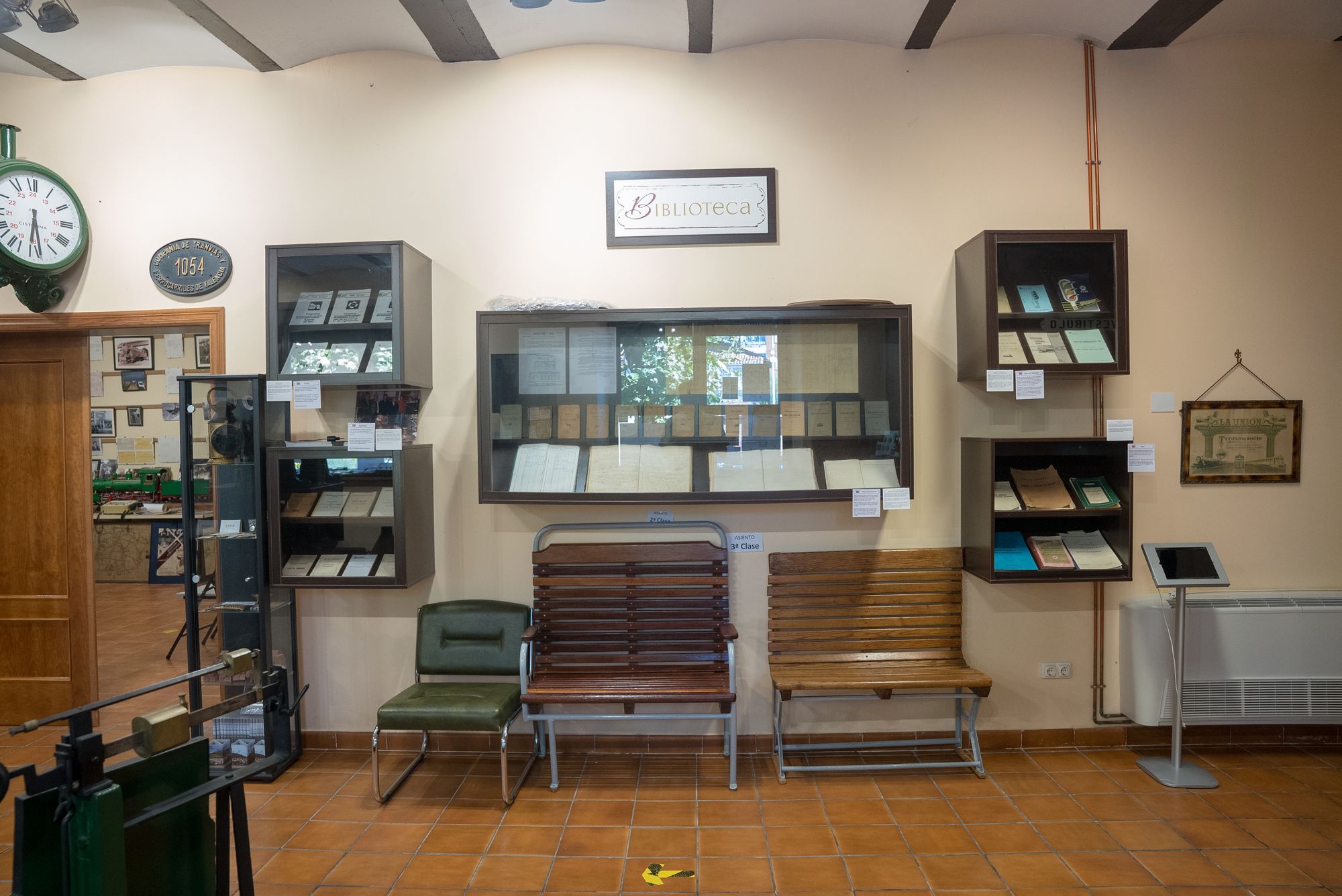 Vitrina Biblioteca con manuales y tarifas, tres asientos y reloj de pared.