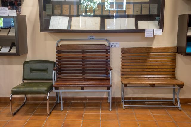 Detalle horizontal de tres asientos: tapizado verde, banco de listones oscuros y banco de madera clara con patas metálicas; vitrina encima.